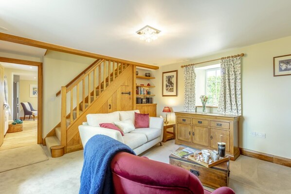 A living room with a wooden staircase, a white sofa, a red armchair, a wooden cabinet, and a window with curtains. - 1 Bed in Blackborough (oc-w39002) (Blackborough)