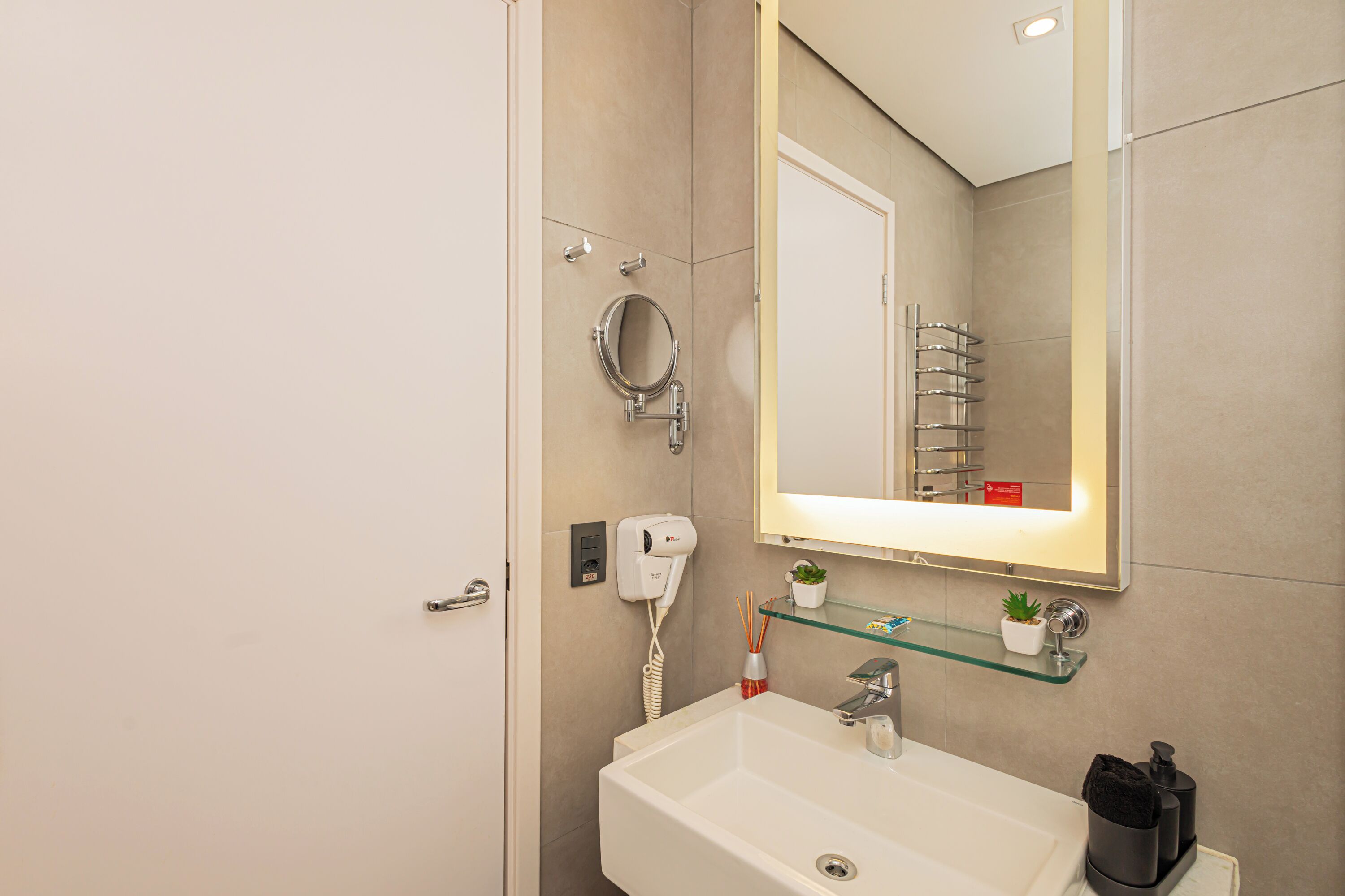 Une salle de bain moderne avec un lavabo blanc, un miroir, un sèche-cheveux et une étagère en verre sur laquelle est posée une plante.