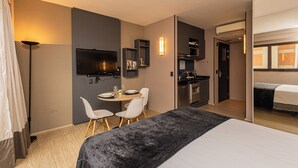 A hotel room with a bed, a dining area with chairs, a TV, a kitchen area, and a door leading to another room.