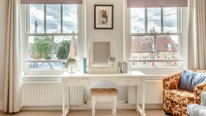 Una habitación con un escritorio blanco, dos ventanas con vistas, un espejo y un sofá.