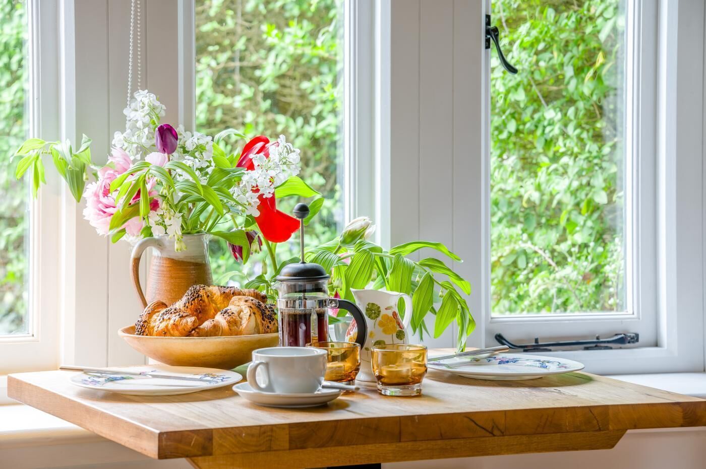 Ontbijt met croissants, een koffiepot, een kopje en een bloevas bij een raam met groen buiten.
