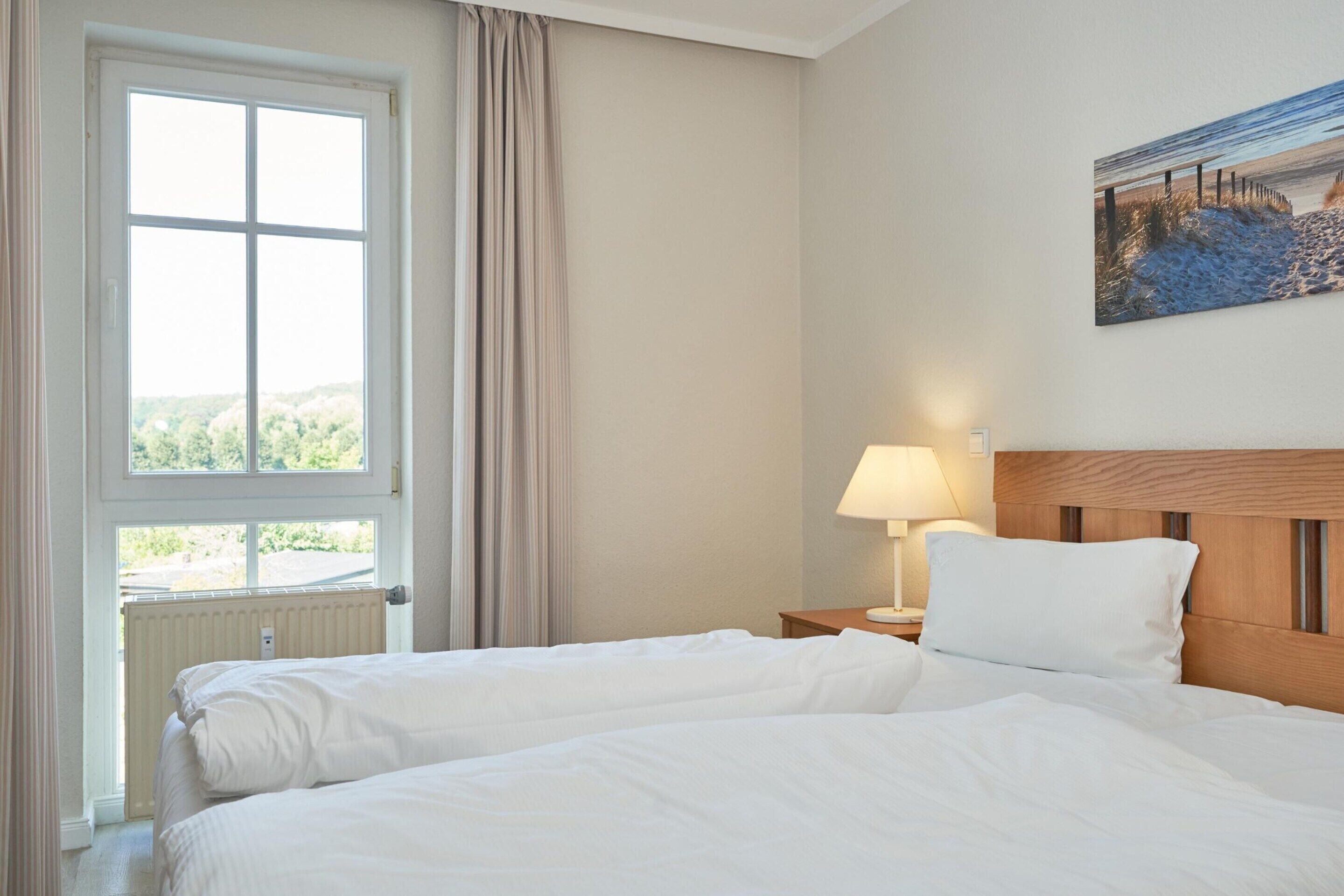 A hotel room with a large window, a bed, a nightstand with a lamp, and a painting on the wall.