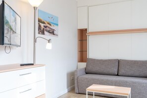 A modern room with a grey sofa, a small wooden table, a TV on a white cabinet, and a lamp. - Fantastisches Studio im Seepark Sellin mit Balkon (Sellin)