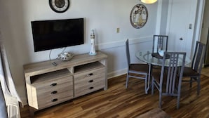 Quarto com televisão de tela plana, armário de madeira, mesa de jantar com cadeiras e relógios de parede.