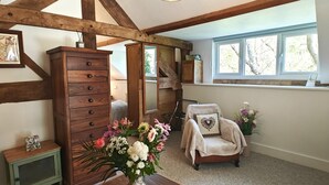 Ein Zimmer mit freiliegenden Holzbalken, einem Holzschrank, einem Stuhl mit Kissen in Herzmuster und einem Fenster mit Blick auf Bäume.