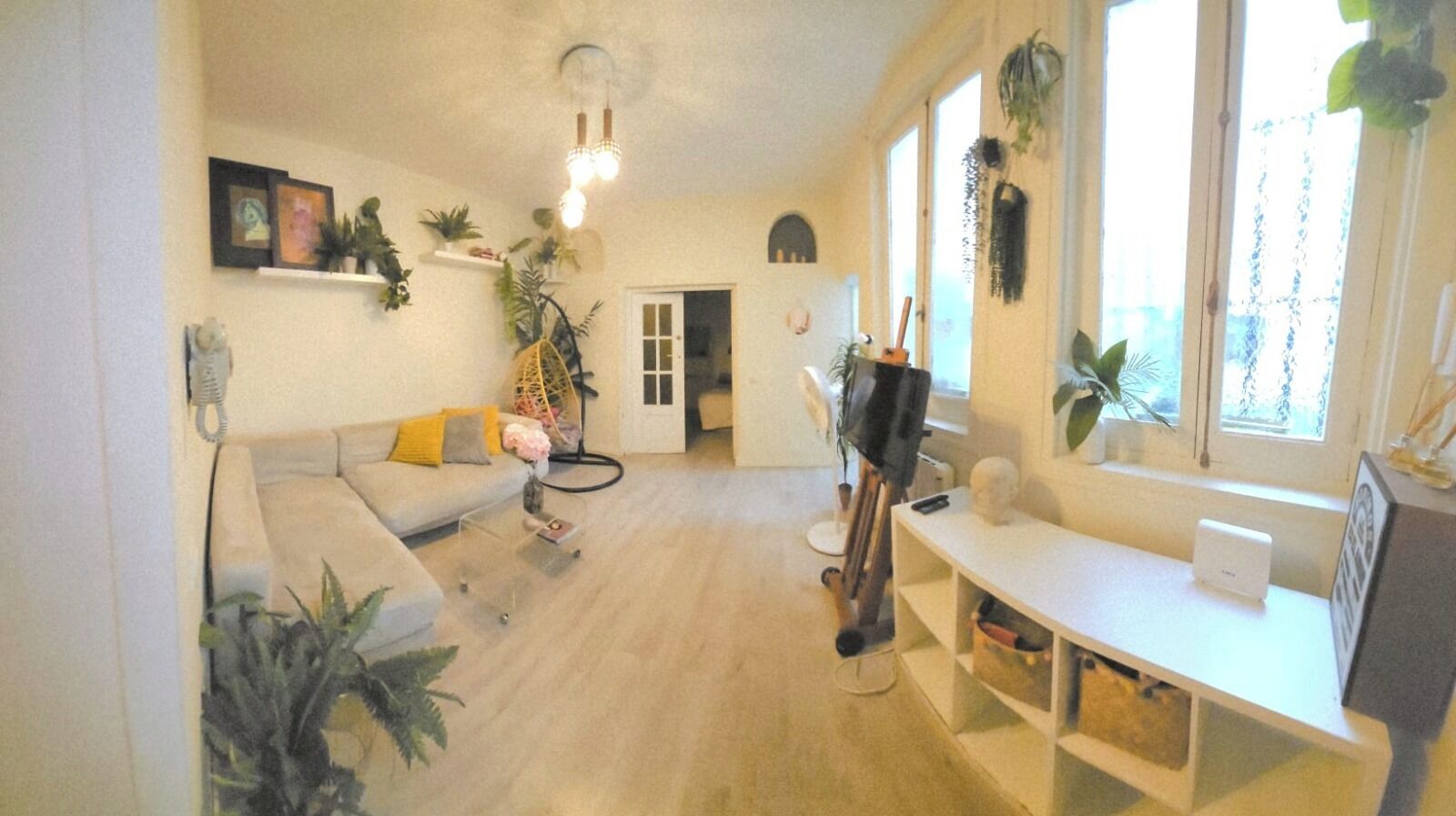 Een woonkamer met een bank, een salontafel, een tv-meubel en een bureau. Er staan planten en er is een hanglamp.