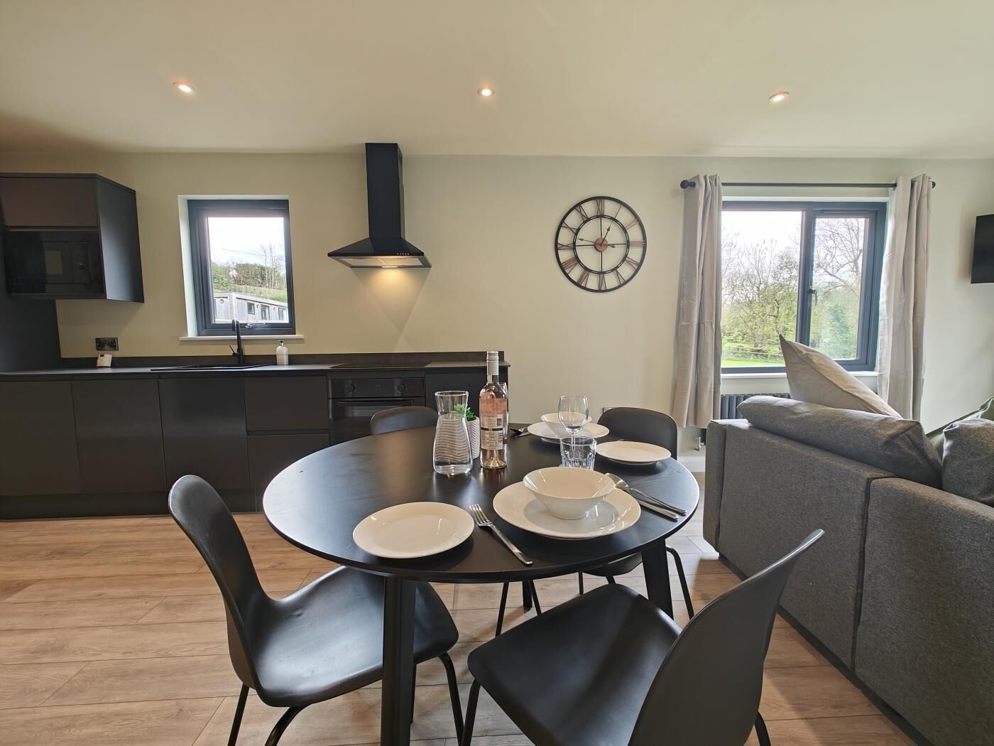 Une cuisine moderne avec des meubles de couleur sombre, une table de salle à manger ronde entourée de chaises, un four encastré et une grande fenêtre.