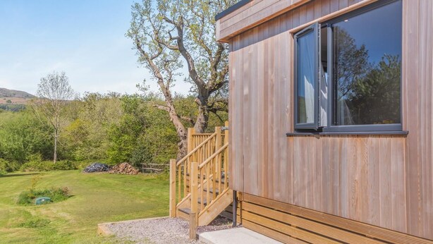 A wooden cabin with a staircase leading to a deck, surrounded by a grassy yard and trees. - 2 Bed in Tyn-y-Groes (oc-w42450) (Ty'n-y-groes)