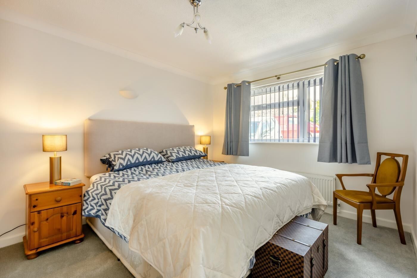 A bedroom with a bed, bedside lamps, a chair, a window with curtains, and a radiator.