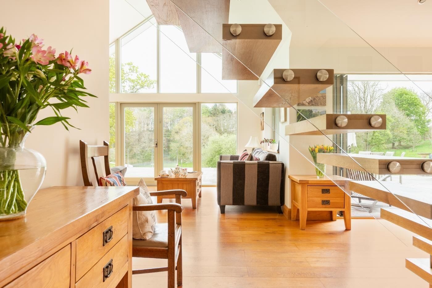 A modern kitchen with wooden cabinets, a dining area with a striped sofa, and large windows offering a view of greenery.