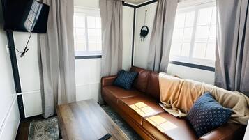 Una sala de estar con un sofá de cuero marrón, una mesa de centro de madera, un televisor montado en la pared y una ventana con cortinas.
