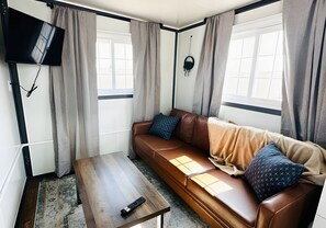 A living room with a brown leather sofa, a wooden coffee table, a TV mounted on the wall, and a window with curtains. - Two Private Bedrooms with Queen and Bunk Beds, Full Kitchen and In-Unit Laundry (Pecos)