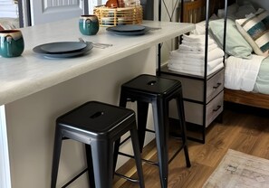 A modern kitchen with a marble countertop, two black stools, a bed with white linens, and a wooden floor.