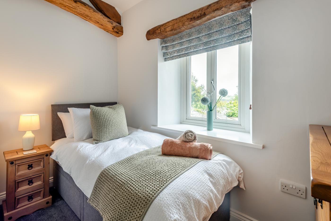 A bedroom with a bed, a nightstand, a window with a view, and a wooden ceiling beam.