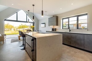 A modern kitchen with a large island, dark cabinetry, and pendant lighting. - 5 Bed in Bluntisham (oc-m40249) (Bluntisham)
