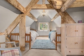 A bedroom with exposed wooden beams, a bed with white bedding, a blue rug, a window with curtains, and a wooden wardrobe.