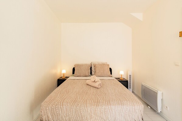 A bedroom with a bed, two bedside tables, two lamps, a radiator, and a window.