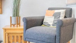 Un fauteuil gris avec un coussin, une petite table d’appoint en bois et une grande plante dans un vase.