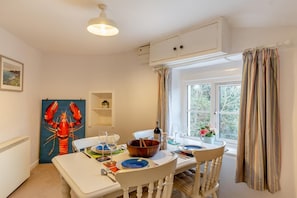 Un coin repas avec une table dressée pour deux personnes, un tableau représentant un homard et une fenêtre avec des rideaux.