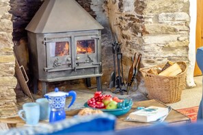 Une cheminée en pierre avec un poêle en métal, un panier de bois de chauffage et une table sur laquelle se trouvent une tasse, une assiette de fruits et un livre.