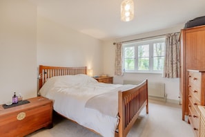 Ein Schlafzimmer mit einem hölzernen Bett, Nachttischen, einer Kommode, einem Fenster mit Vorhängen und einem Heizkörper.