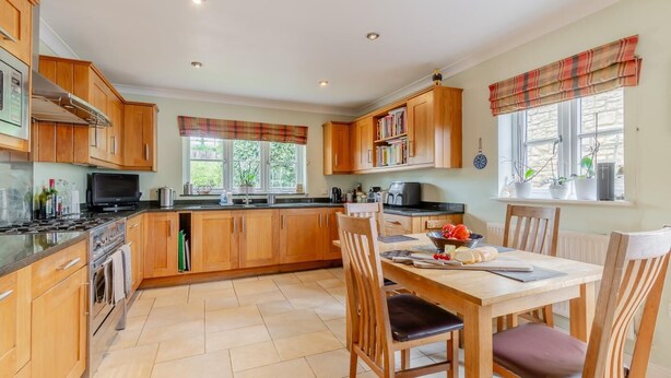 A kitchen with wooden cabinets, a dining table with chairs, and a window with a view of greenery. - 4 Bed in Northleach (oc-b42441) (Northleach)