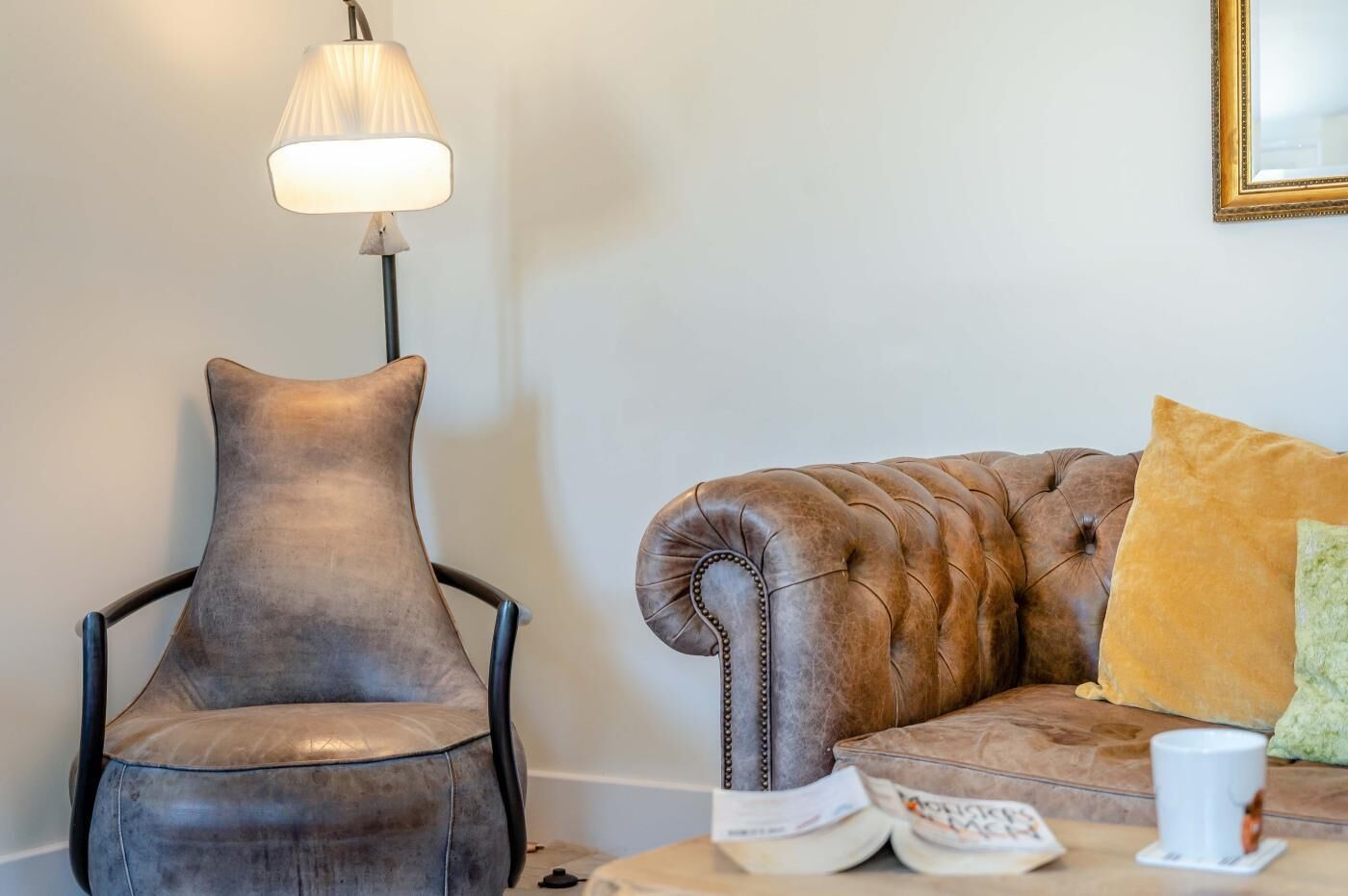 A room with a leather armchair, a tufted sofa with a cushion, a floor lamp, and a remote control on a table.