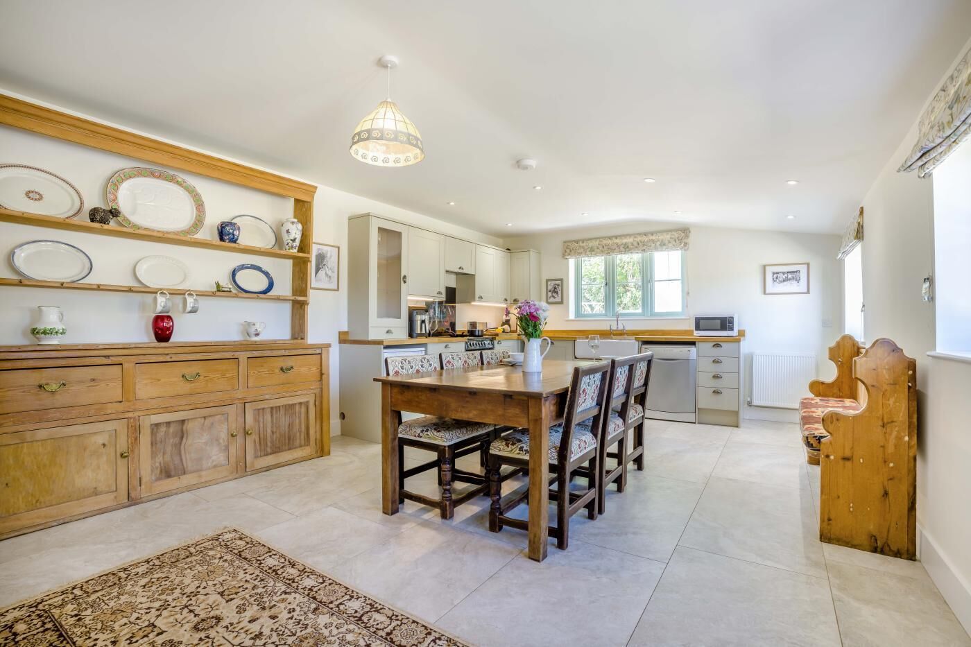 A dining area with a wooden table and chairs, a kitchen in the background, and a patterned rug on the floor.