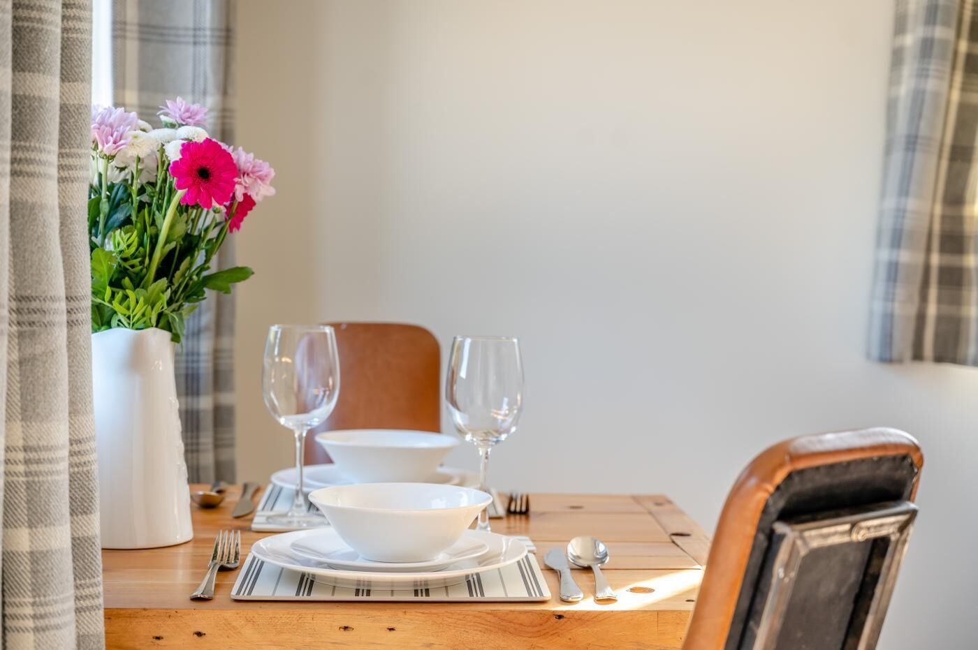 A dining table set with a white plate, two wine glasses, a bowl, and cutlery. A vase with flowers is on the table.