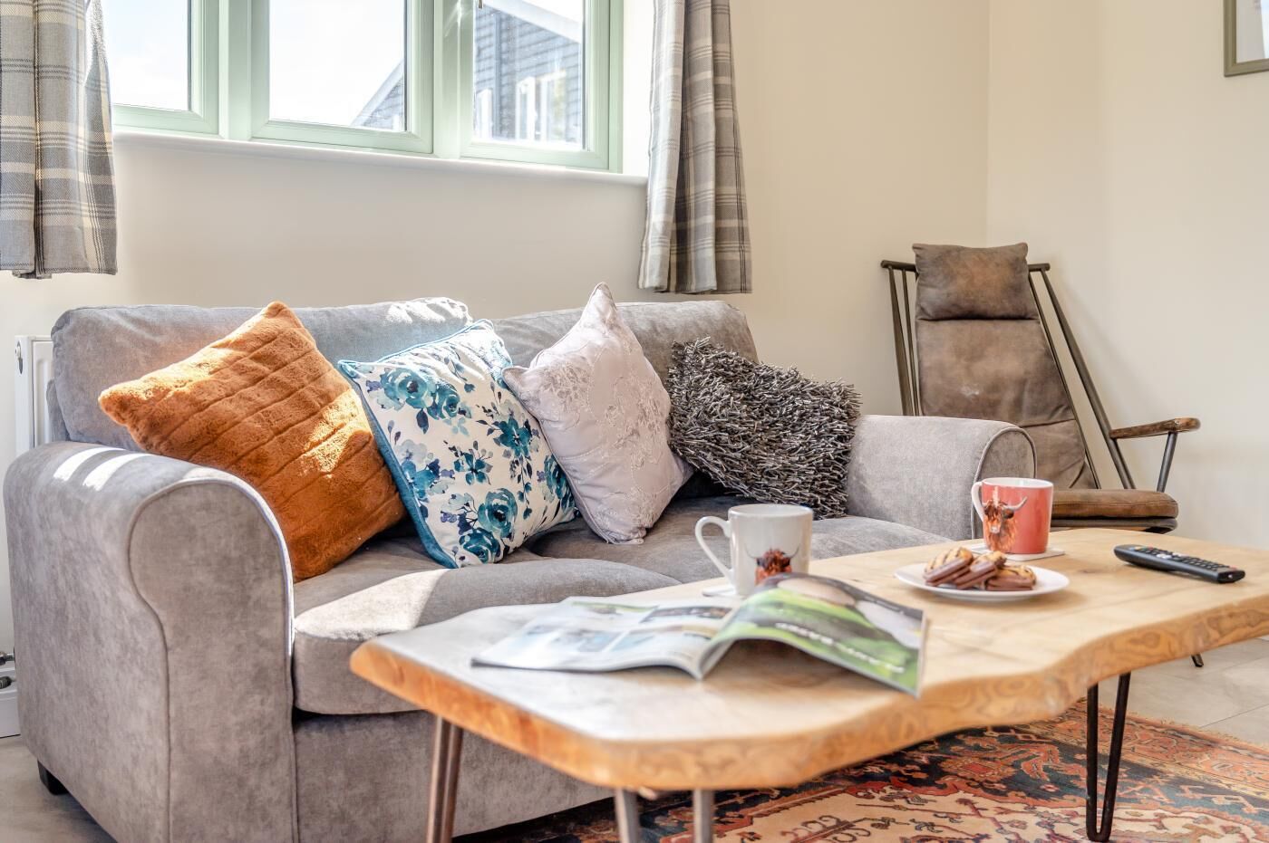 A living room with a grey sofa, multiple cushions, a coffee table with a magazine, a mug, and a remote control.