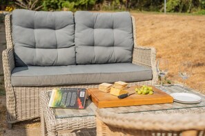 A wicker sofa with grey cushions, a book titled 'Angels,' a wooden board with cheese and grapes, and two wine glasses on a glass table. - 1 Bed in Brockenhurst (oc-f40681) (Brockenhurst)