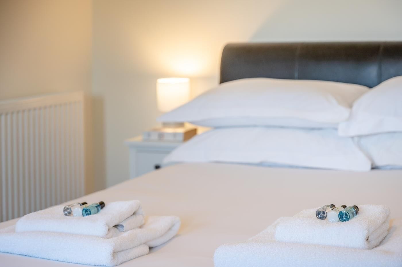 Una cama bien hecha con sábanas y toallas blancas, una mesita de noche con lámpara, y un radiador al fondo.