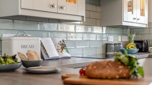 A kitchen with a tiled backsplash, a bread box, a cookbook, a bowl of salad, and a loaf of bread on a cutting board. - 2 Bed in St. Merryn (oc-b14684) (Saint Merryn)