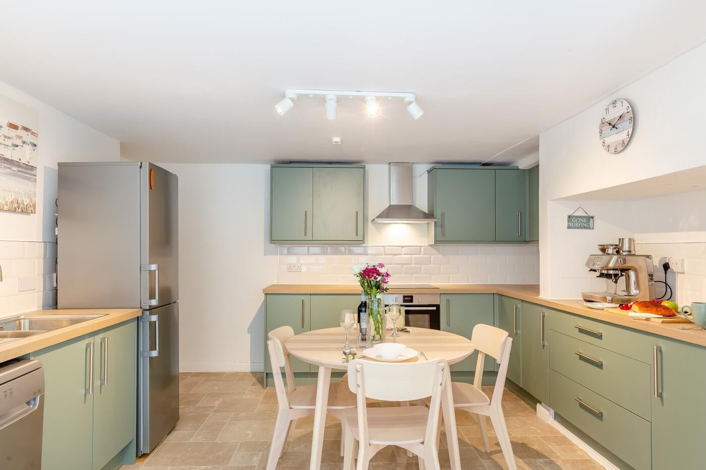 A modern kitchen with teal cabinets, a stainless steel refrigerator, a round dining table with chairs, and a coffee machine.