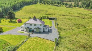Une grande maison avec une allée pavée et une cour clôturée, entourée de champs verdoyants et de montagnes.