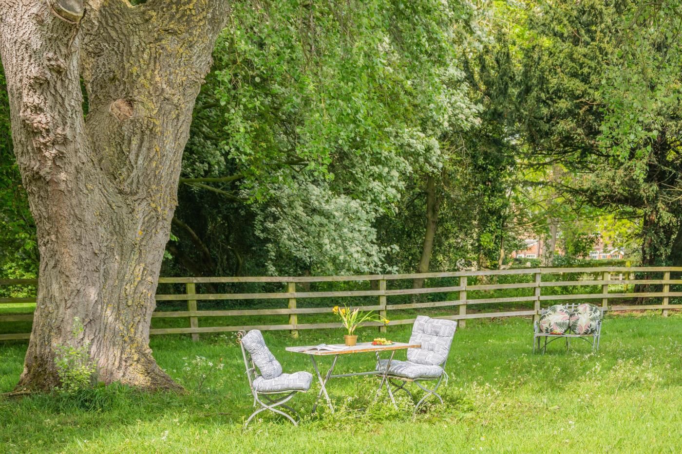 A rustic outdoor seating area with two chairs and a table, surrounded by lush greenery and a wooden fence.