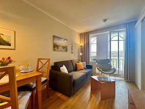 Ein Wohnzimmer mit einer Couch, einem Esstisch und einem Stuhl. Ein Fenster mit Vorhängen bietet Blick ins Freie.