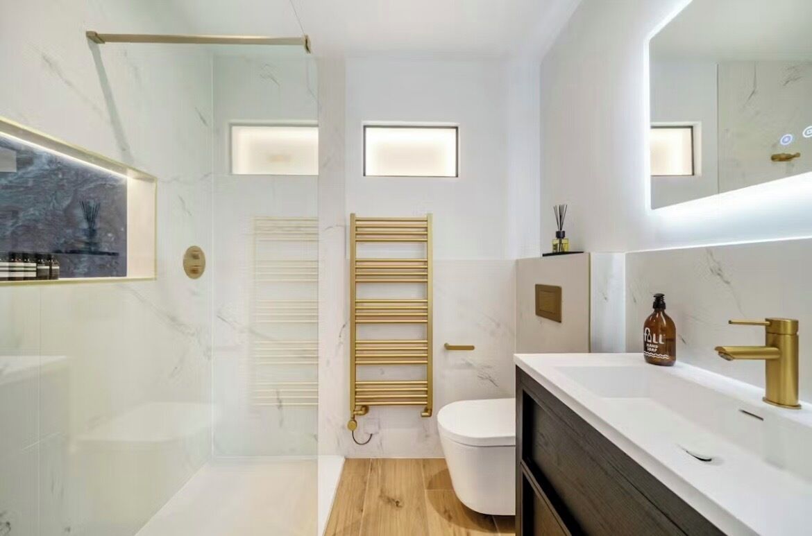 A modern bathroom with a glass shower enclosure, a white toilet, a gold towel rack, and a white sink with gold fixtures.