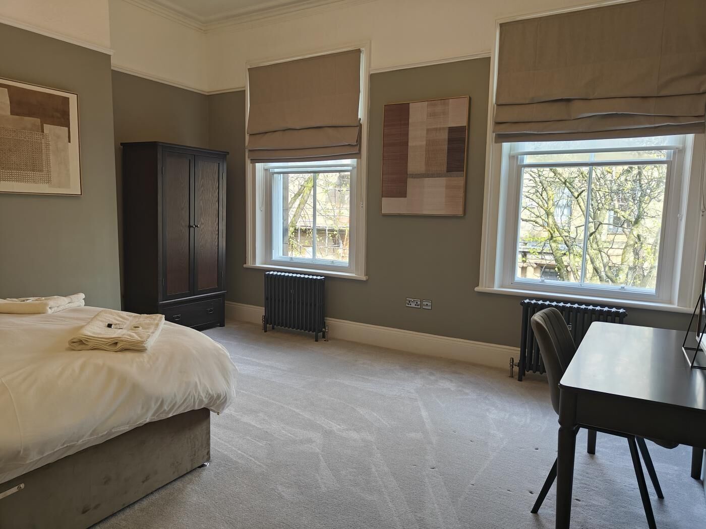 A bedroom with a bed, a desk, a radiator, and large windows.
