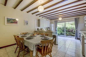 A dining area with a table set for four, wooden chairs, a kitchen in the background, and a view of greenery through the glass door. - 3 Bed in Stackpole (oc-c41424) (Stackpole)