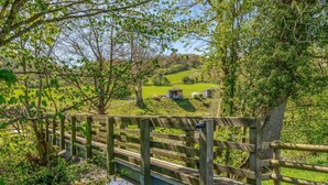 A wooden bridge over a body of water, surrounded by lush greenery and a rural landscape with farm buildings. - 1 Bed in Aberbechan (oc-t41392) (Newtown)