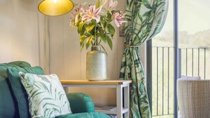 A room with a teal sofa, a small table with a vase of flowers, a lamp, and a view through a window with curtains. - 1 Bed in Aberbechan (oc-t41392) (Newtown)