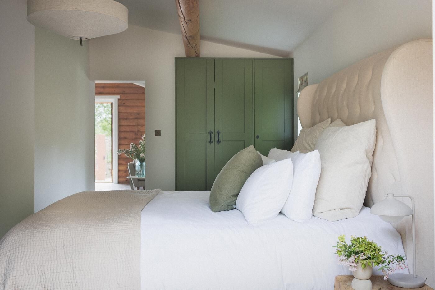 A bedroom with a bed, pillows, a bedside table with a lamp, a green wardrobe, and a window.