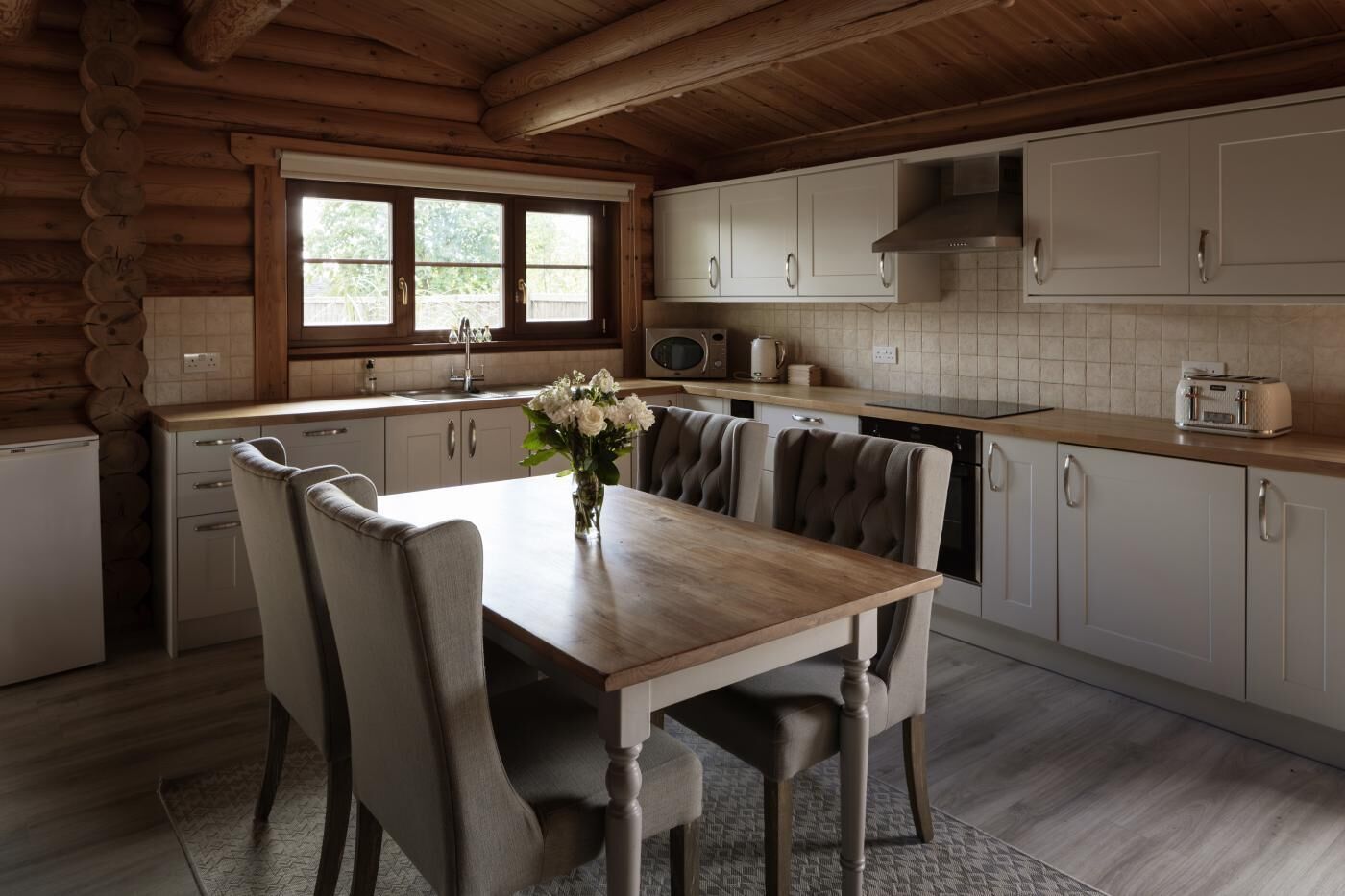 Une cuisine rustique aménagée dans une cabane en rondins, avec une table à manger, des chaises, un vase de fleurs et une fenêtre.