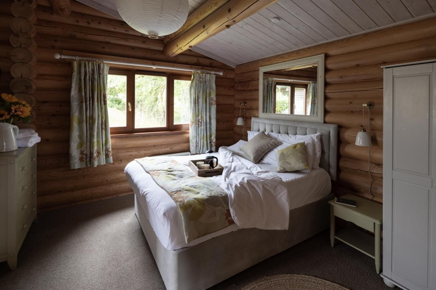 Une chambre à coucher située dans une cabane en rondins, avec un lit, des rideaux, une table de chevet et une commode.