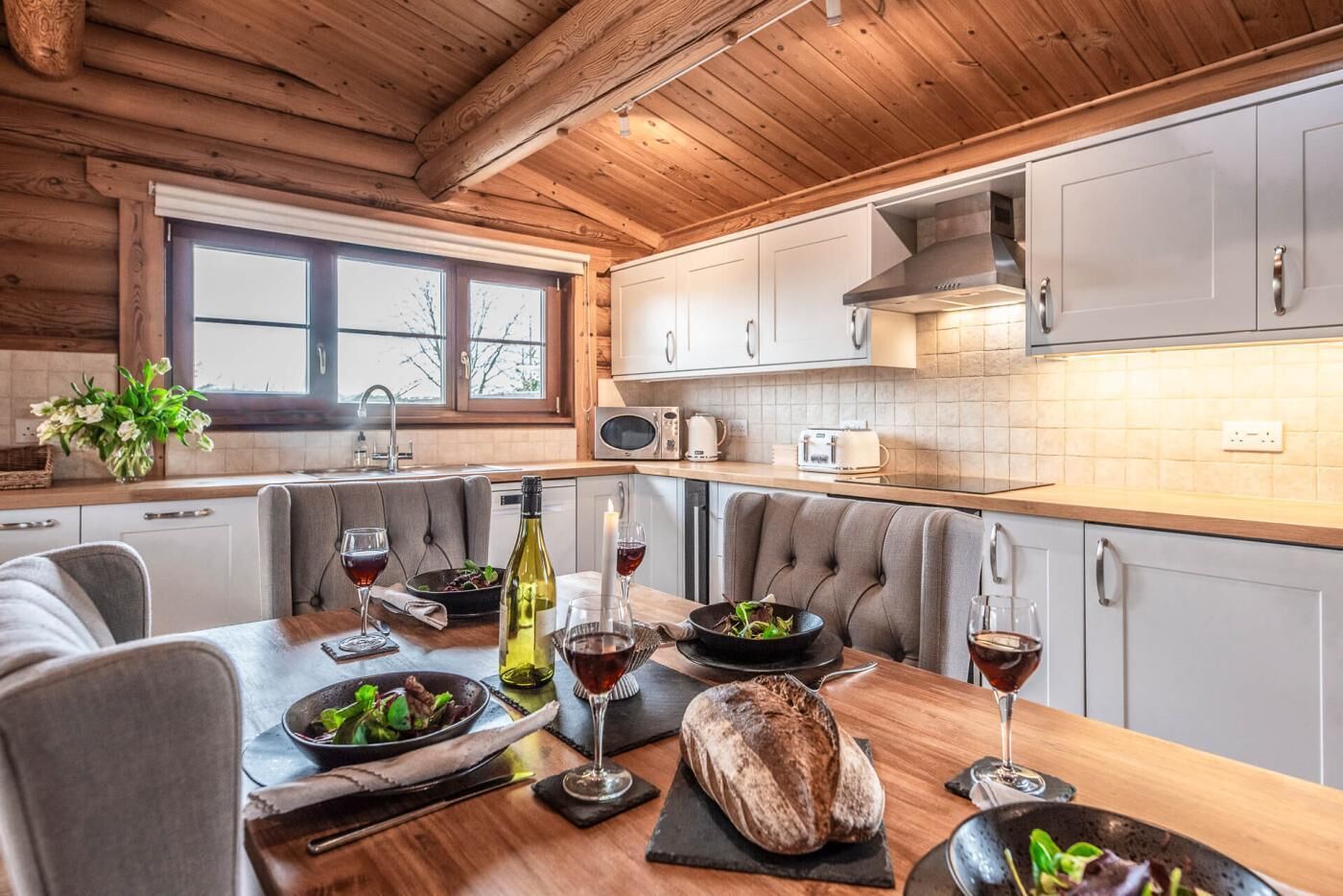 Une cuisine moderne avec des poutres en bois au plafond, des meubles blancs et un coin repas doté de chaises grises recouvertes de tissu.