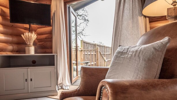 A wooden interior with a leather sofa, a flat-screen TV, and a view of a wooden deck outside.