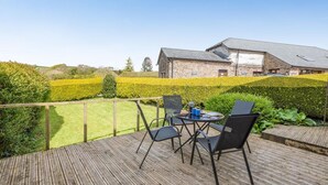 Une terrasse en bois avec des sièges extérieurs, un bâtiment en pierre en arrière-plan et des haies bien entretenues.
