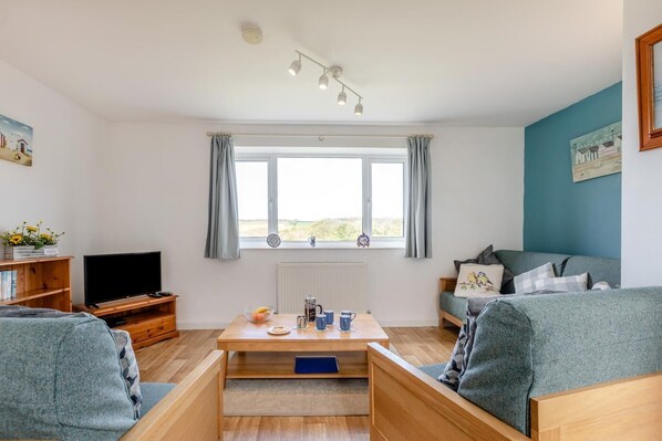 A living room with a TV, wooden furniture, a window with curtains, and a view of the outdoors. - 2 Bed in Bideford (oc-73954) (Hartland)