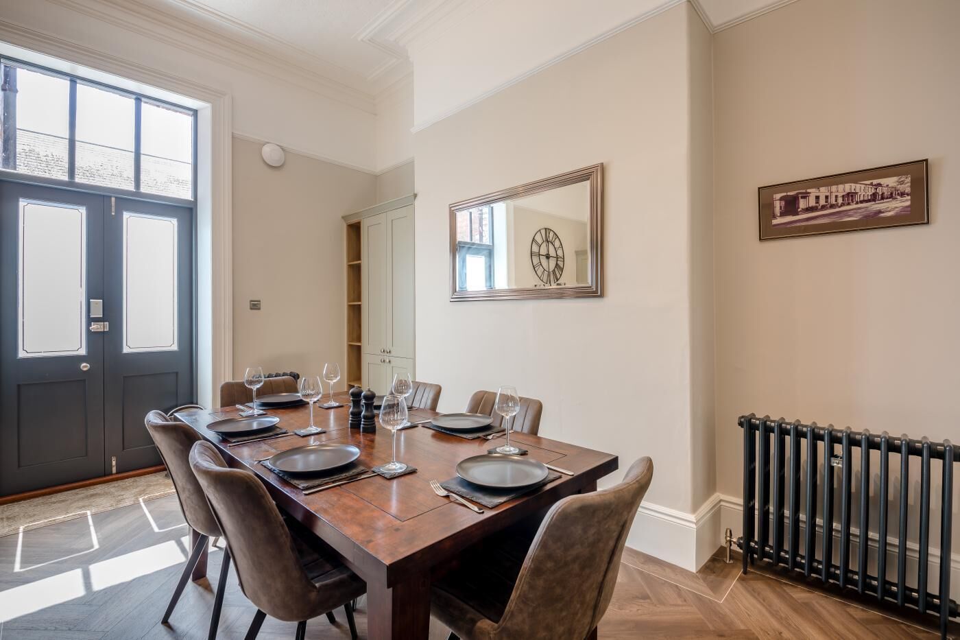 A dining room with a large table set for six, a black door, a clock on the wall, and a radiator.